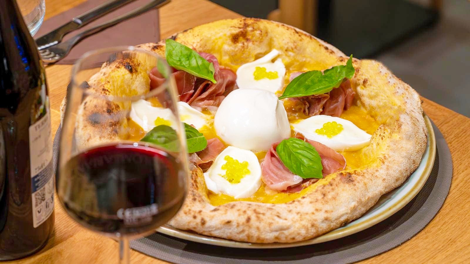 La Cortile Abatellis: base di datterino giallo, una burrata intera al centro, quenelle bianche con sopra un ciuffo di perle gialle di olio extravergine, falde di prosciutto crudo e foglie di basilico, con un calice di vino rosso in primo piano.