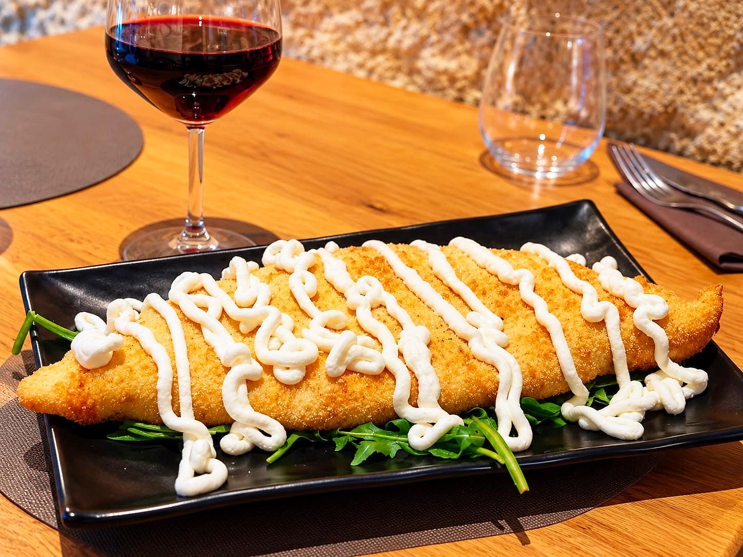 Il Calzone Campano su un piatto nero rettangolare: la sfoglia dorata e croccante attraversata da strisce di crema di ricotta, su un letto di rucola, con un calice di rosso e il muro di tufo dietro.
