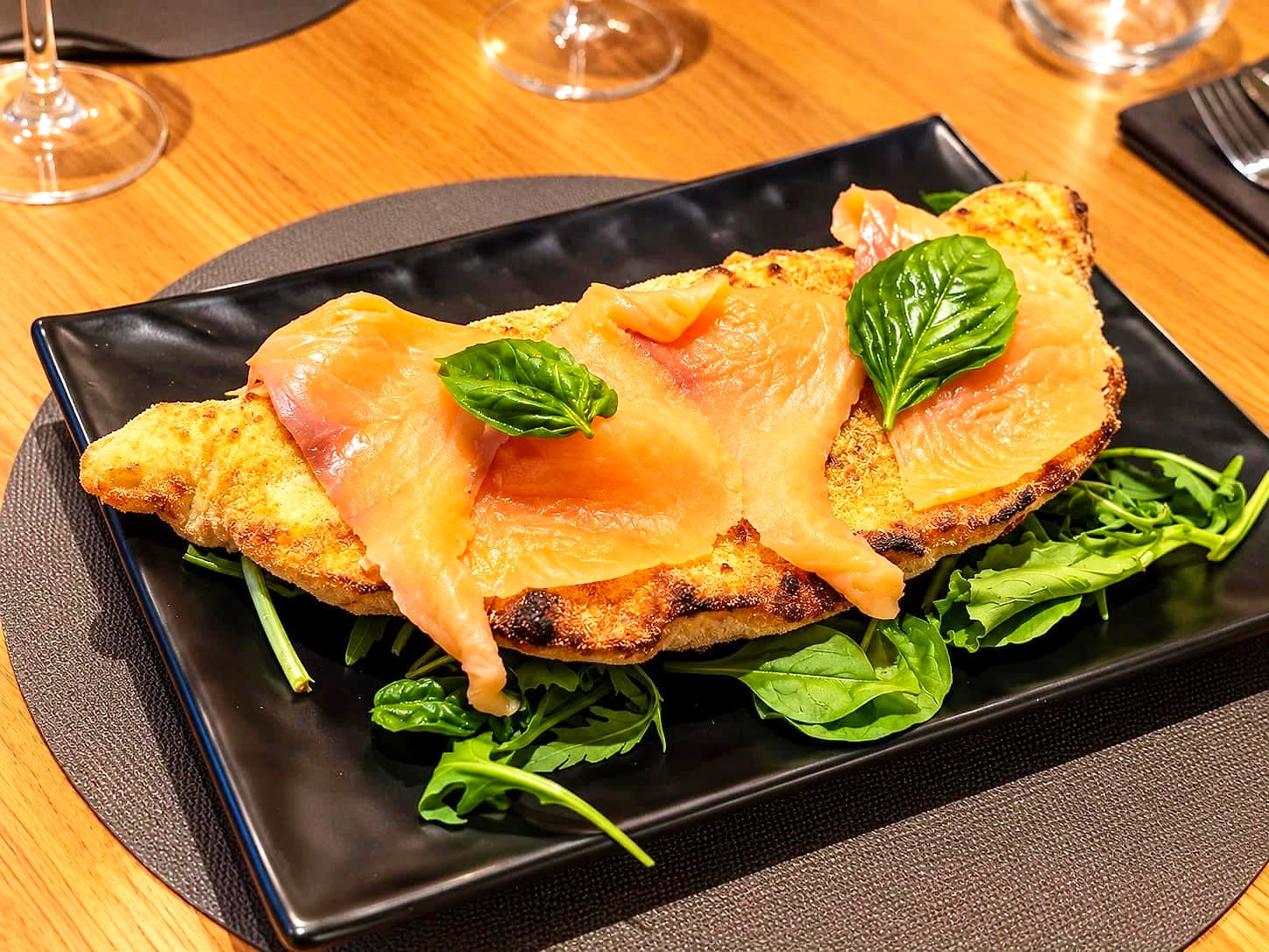 Il Calzone Premium su un piatto nero rettangolare: la sfoglia dorata e croccante coperta di fette di salmone affumicato e foglie di basilico, su un letto di songino e rucola, con i calici sul tavolo di rovere.