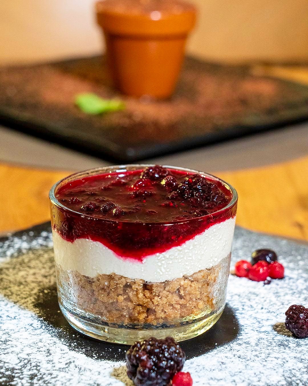 Cheesecake ai frutti di bosco servita nel bicchiere, con la salsa di frutti rossi sopra, su una lastra di ardesia spolverata di zucchero a velo.
