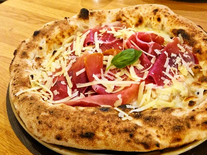 Pizza bianca con prosciutto crudo e scaglie di formaggio stagionato, cornicione alto e maculato dalla cottura.