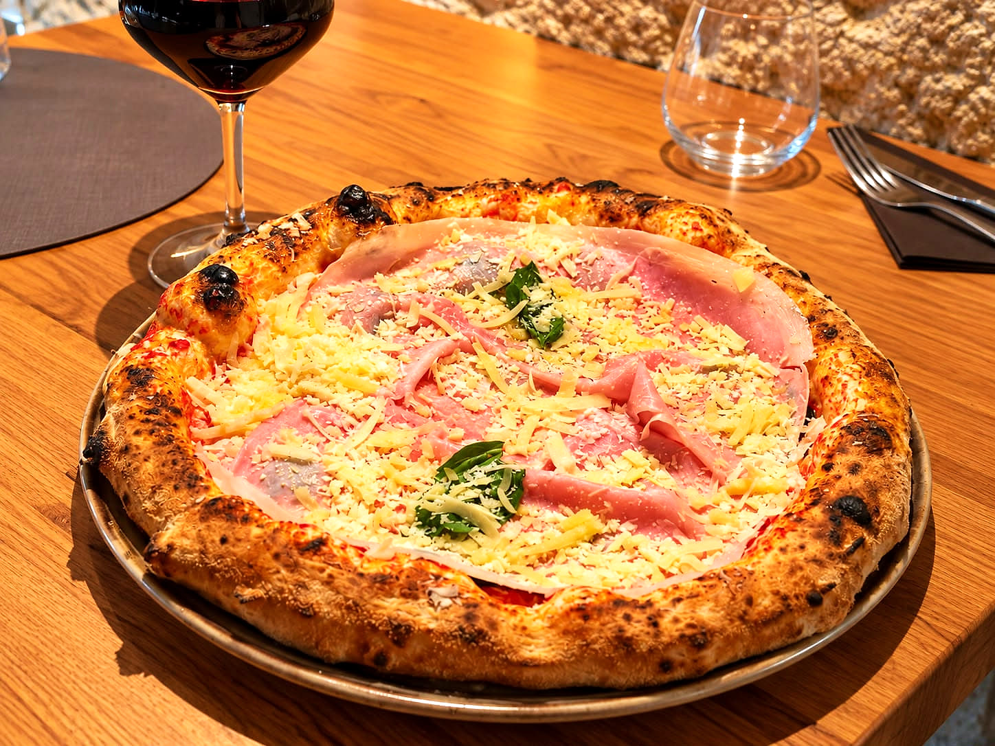 La Greca: pomodoro, fiordilatte, le fette di melanzane fritte sotto le falde di prosciutto cotto, il caciocavallo grattugiato e due ciuffi di basilico, su un cornicione alto e bruciacchiato, con un calice di rosso sul tavolo di rovere.