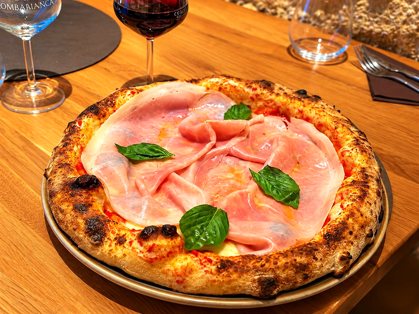 La Romana: pomodoro, fiordilatte, falde di prosciutto cotto e foglie di basilico su un cornicione alto e bruciacchiato dalla fiamma, con i calici di vino e d'acqua sul tavolo di rovere.