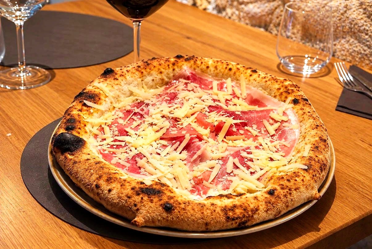 La Cortile Sambuca sul tavolo di rovere: falde di prosciutto crudo di Parma e scaglie di Parmigiano Reggiano sulla bufala, su un cornicione alto e maculato dalla cottura, con un calice di rosso e il muro di tufo dietro.