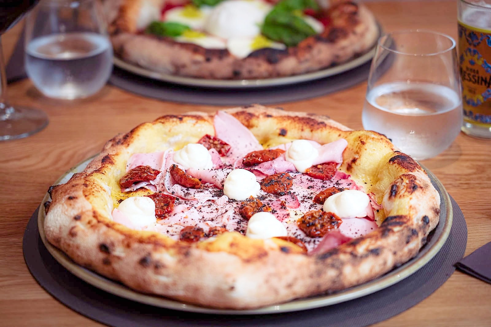 La Cortile Sclafani su un piatto di ceramica, sul tavolo di rovere: falde di prosciutto cotto affumicato, ciuffi di ricotta fresca, datterini rossi secchi e una spolverata di polvere di olive nere, su un cornicione alto e maculato dalla cottura. Dietro, sfocati, una seconda pizza, i bicchieri d'acqua e un calice di Birra Messina.