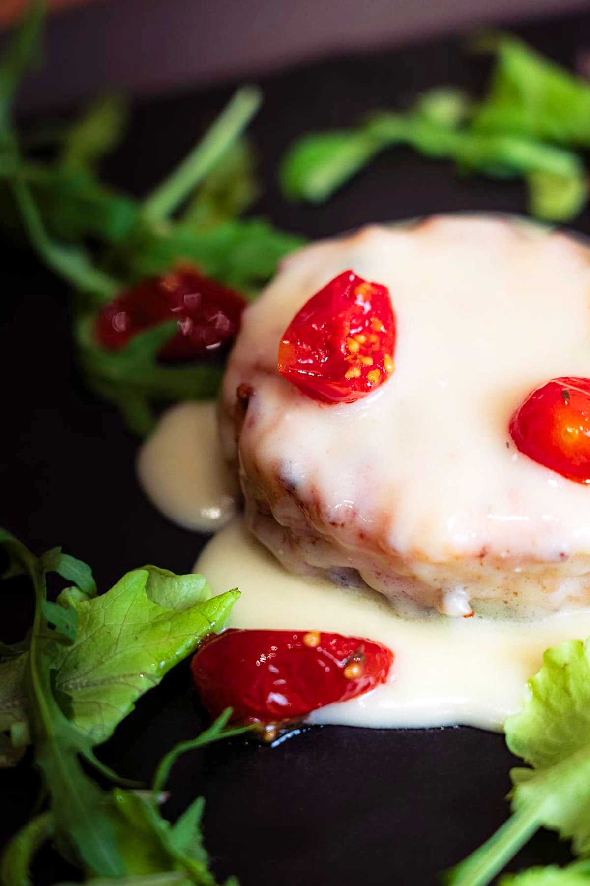 Il timballo di pasta croccante su una lastra nera, coperto da una crema chiara che cola lungo i bordi, con i pomodorini confit sopra e l'insalatina tutt'intorno.