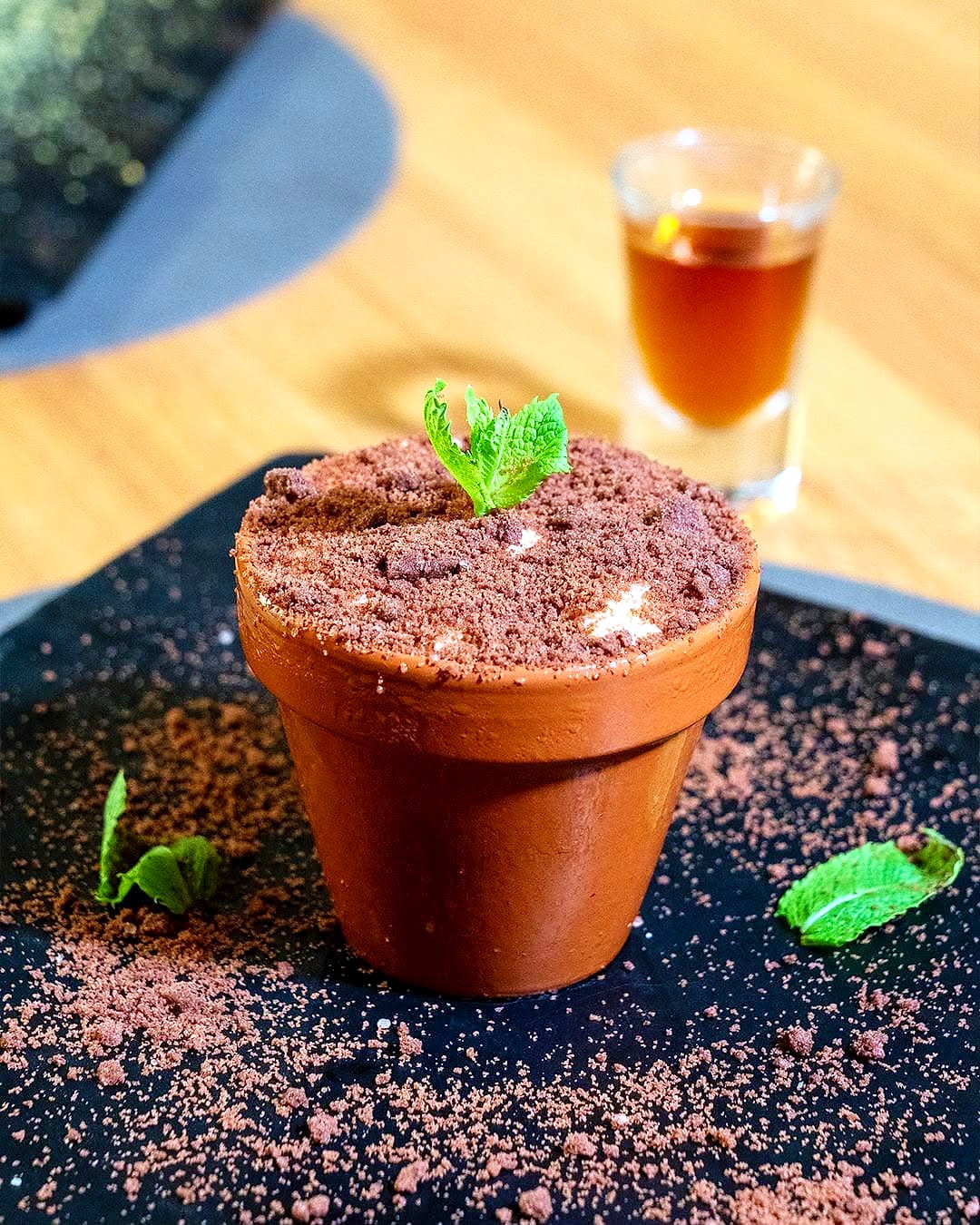 Tiramisù servito in un vasetto di terracotta, coperto di cacao come fosse terra, con una foglia di menta e un bicchierino di liquore accanto.