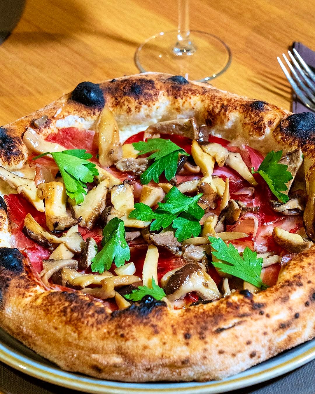Pizza su base bianca di crema di pecorino coperta di funghi cardoncelli saltati, con il capocollo sotto e ciuffi di prezzemolo fresco sopra, servita su un piatto dal bordo dorato.