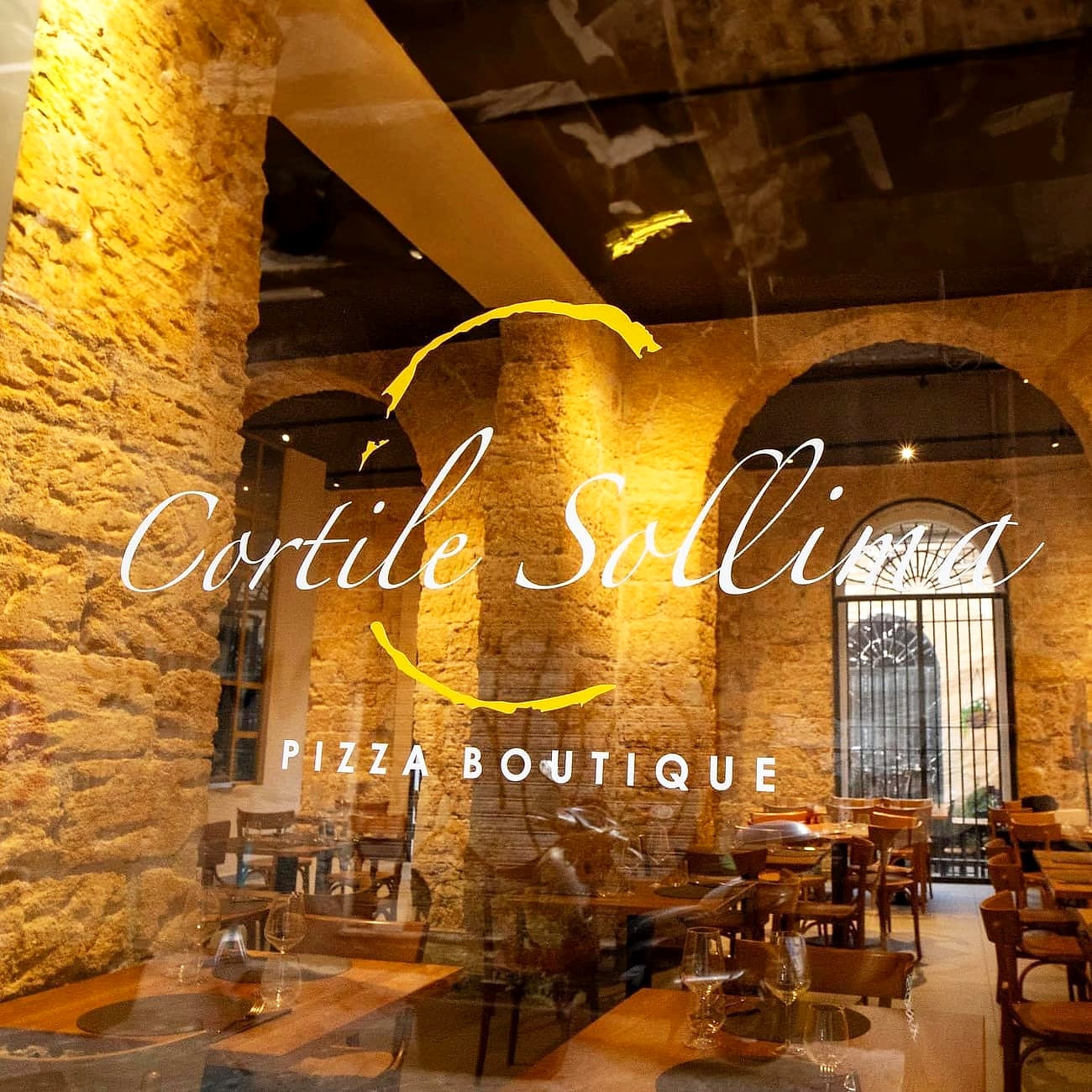 La vetrata di via Calascibetta di sera: il nome Cortile Sollima inciso in bianco sul vetro, dentro un arco di pennello dorato, con sotto la scritta PIZZA BOUTIQUE, e dietro il vetro i pilastri e le arcate di tufo illuminati, la finestra ad arco con la grata e i tavoli apparecchiati.